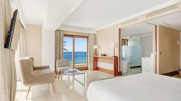 Ein Hotelzimmer mit einem großen Bett, einem Sessel, einem Tisch, einem Schreibtisch, einem Spiegel und Meerblick.