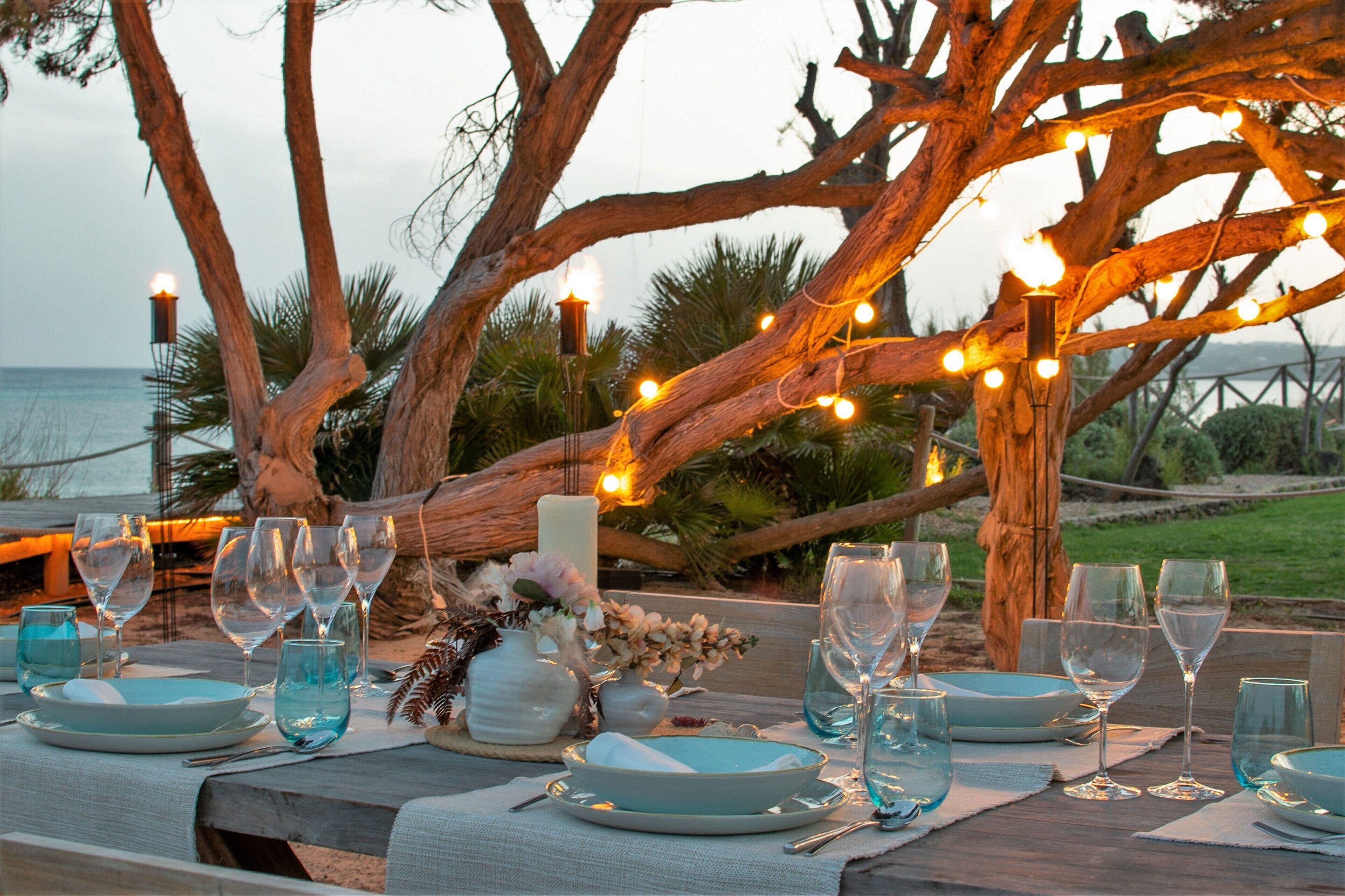 A table set with dinnerware and glasses, decorated with candles and string lights, under a large tree.