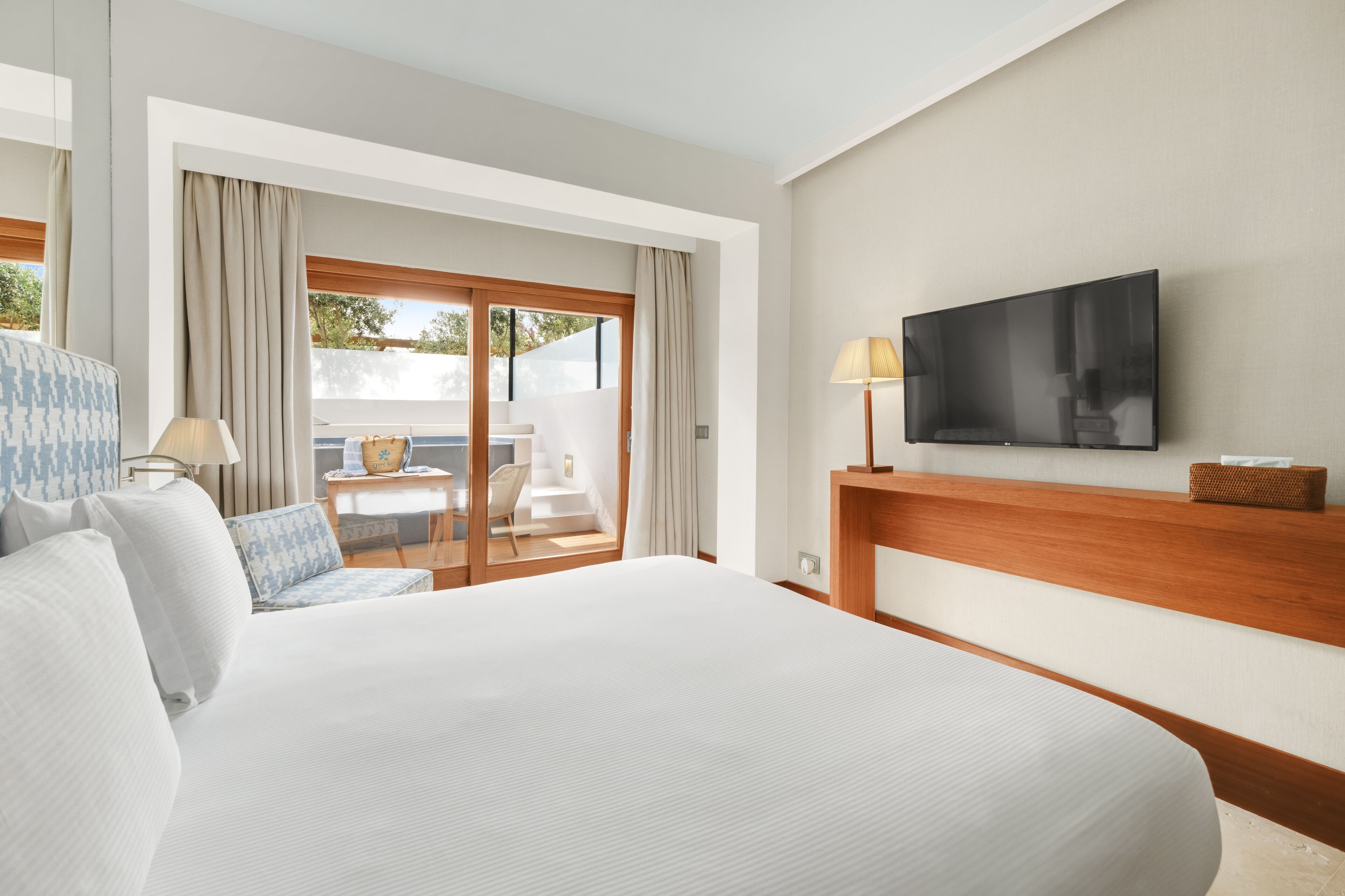 Ein Hotelzimmer mit einem großen Bett, einem an der Wand befestigten Fernseher, einem Schreibtisch mit Lampe und Blick ins Freie durch eine Schiebetür.