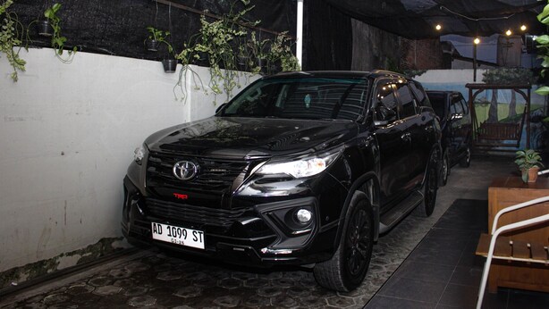 A black SUV parked in a covered outdoor area with a tiled floor, surrounded by greenery and a rustic wooden bench. - Capital O 2258 Santana Syariah Guest House (Surakarta)