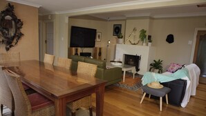 Ein Essbereich mit einem Holztisch, Korbstühlen, einem grünen Sofa, einem Fernseher und einem Kamin.