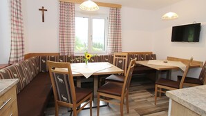 A dining area with a wooden table, chairs, and a bench. There is a cross on the wall, a window with curtains, and a television mounted on the wall.