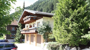 A traditional chalet with a wooden balcony, surrounded by lush greenery and a mountain backdrop.