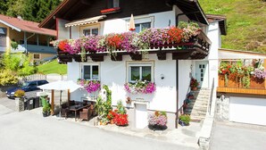 Ein zweistöckiges Gebäude mit einem Balkon, geschmückt mit bunten Blumen, einer kleinen Außen-Sitzgelegenheit und einer Steintreppe zum Eingang.