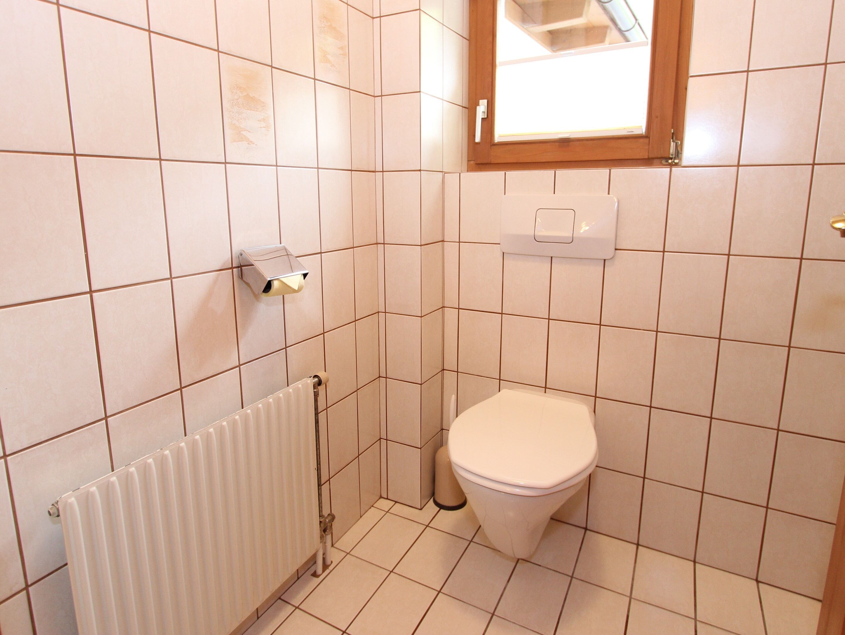 Une petite salle de bain carrelée, équipée de toilettes, d’un radiateur et d’une fenêtre.