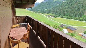 A wooden balcony with a table and chairs, overlooking a green valley and forested hills. - Apartment in Viehhofen Near Ski Lifts (Viehhofen)