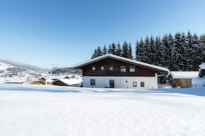 A two-story wooden house with a snow-covered roof, surrounded by a snowy landscape and evergreen trees. - Apartment in Flachau Near Ski Lift (Flachau)