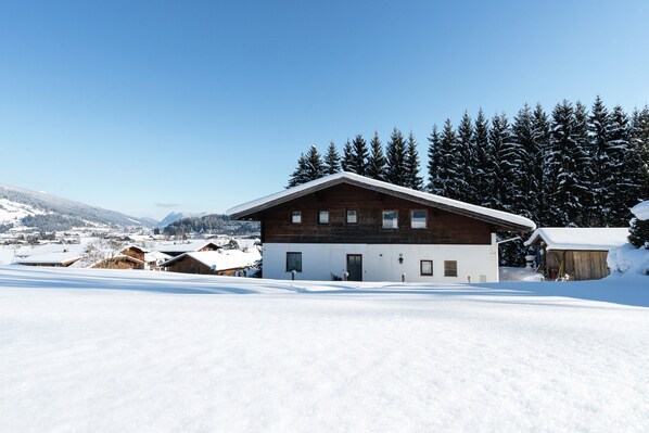 A two-story wooden house with a snow-covered roof, surrounded by a snowy landscape and evergreen trees.