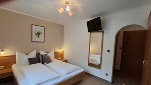 A hotel room with a double bed, bedside tables, a mounted TV, a framed artwork, and a door leading to another room.