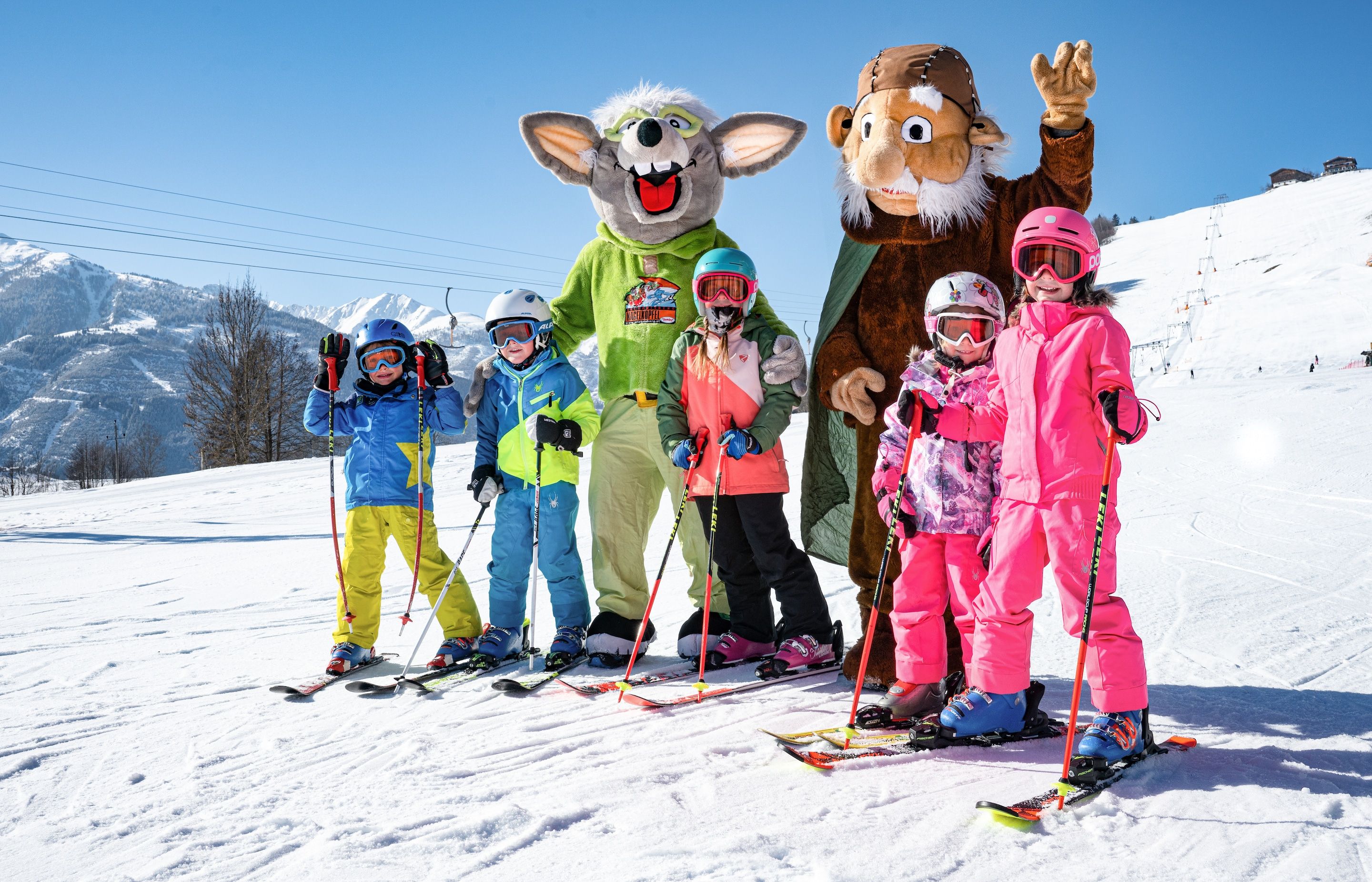 A group of people on a snowy slope with ski equipment, a mascot, and a mountain backdrop.
