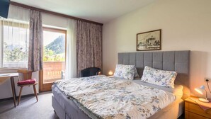 Ein Schlafzimmer mit einem großen Bett, einem Schreibtisch und einem Stuhl. Ein Fenster mit Vorhängen bietet Blick auf Grünflächen.