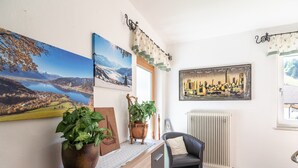 Ein Zimmer mit einem Holztisch, einem Stuhl und einer Heizung. An der Wand hängen drei gerahmte Bilder: eines zeigt eine Stadtsicht, die anderen zwei Landschaftsmotive.