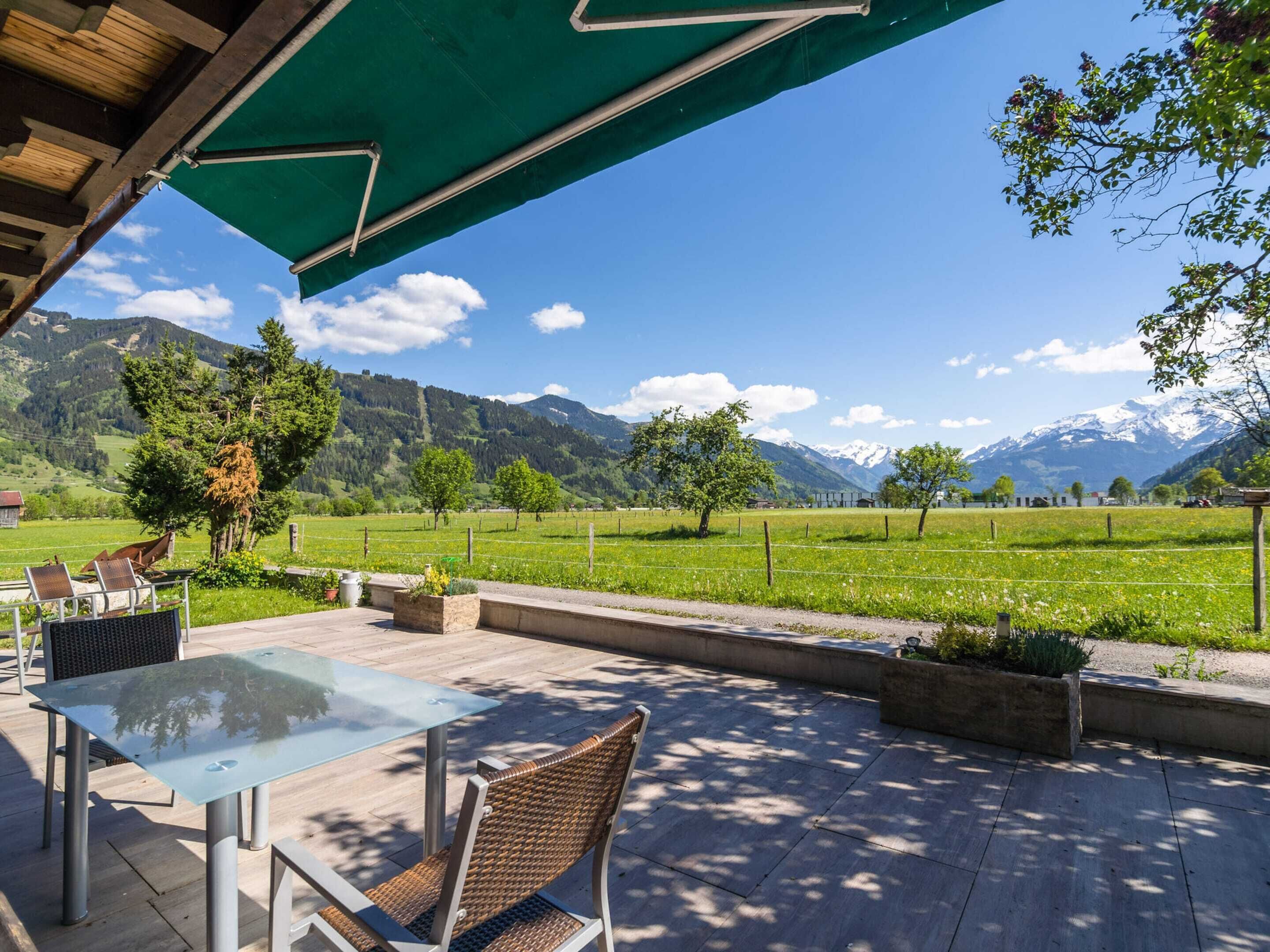 An outdoor patio with a glass table and wicker chairs, overlooking a green field and mountains.