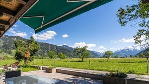 Eine Terrasse im Freien mit einem Glastisch und Korbstühlen, mit Blick auf ein grünes Feld und Berge.