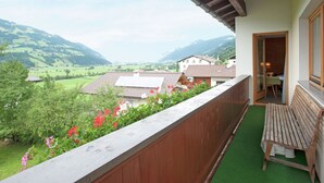 Een balkon met houten hekwerk, een bankje en uitzicht op een vallei met huizen en bergen.