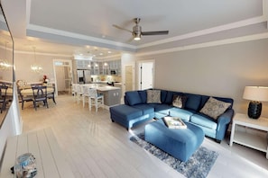 A spacious living room with a blue sectional sofa, a dining area with white chairs, and a kitchen in the background.