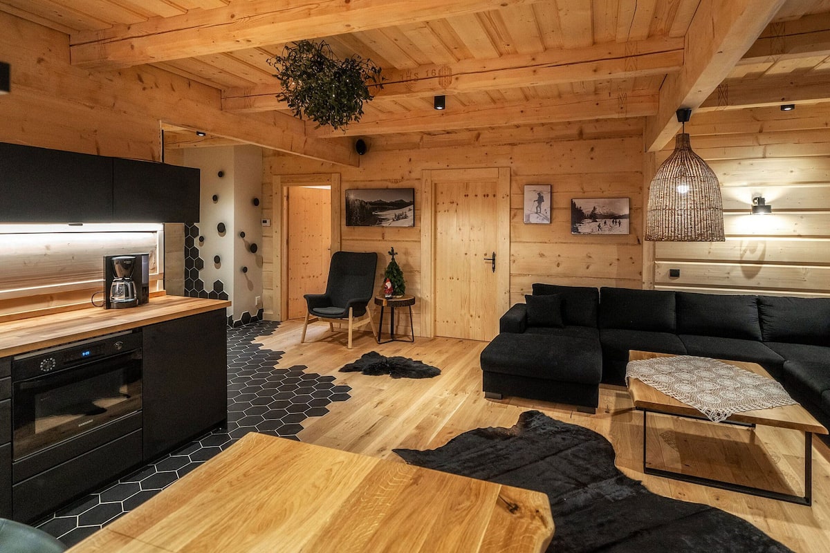 Interior acogedor de una cabaña con techo de madera, una zona de cocina con cafetera, un sofá negro y una pequeña mesa con mantel de encaje.