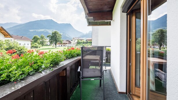 Un balcón con una silla, baranda de madera y vistas a montañas y casas.