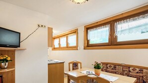Un espacio habitable pequeño con una mesa de comedor, un televisor instalado en la pared, una zona de cocina y una ventana con cortinas de encaje.