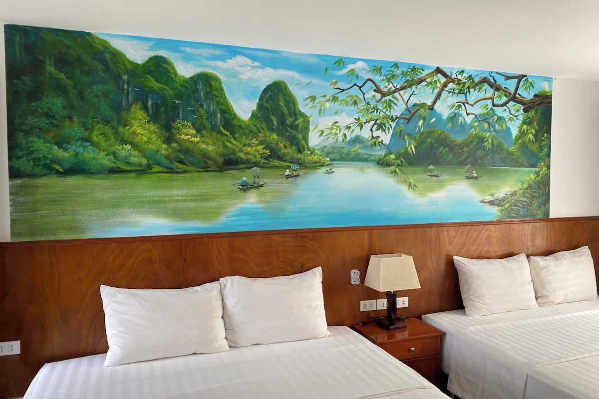 A hotel room with two beds, a nightstand with a lamp, and a large painting of a river landscape on the wall.