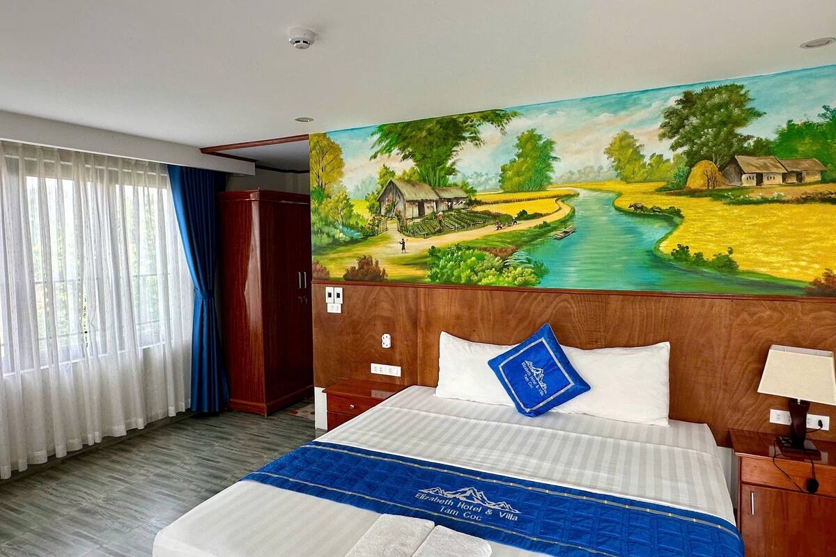 A hotel room with a bed, a painting of a rural landscape, a wooden headboard, and a blue pillow.