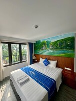 Una habitación de hotel con cama, un cuadro de un paisaje, una ventana con vistas y una mesita de noche.