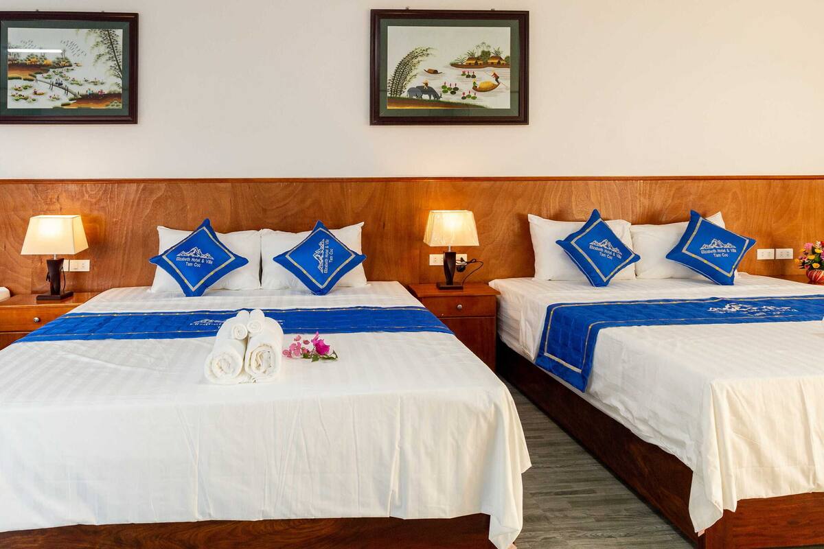 A hotel room with four beds, each with white bedding and blue accents. There are two wall-mounted lamps on wooden nightstands, and the room has a wooden headboard.
