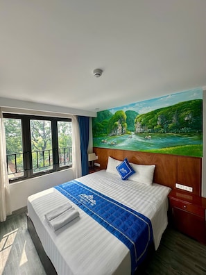 Quarto de hotel com cama, pintura de uma paisagem, janela com vista e mesa de cabeceira.