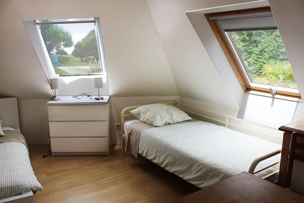 Une petite pièce bien éclairée, avec un plafond en pente, un lit simple, un bureau avec une chaise et une fenêtre donnant sur un paysage verdoyant.