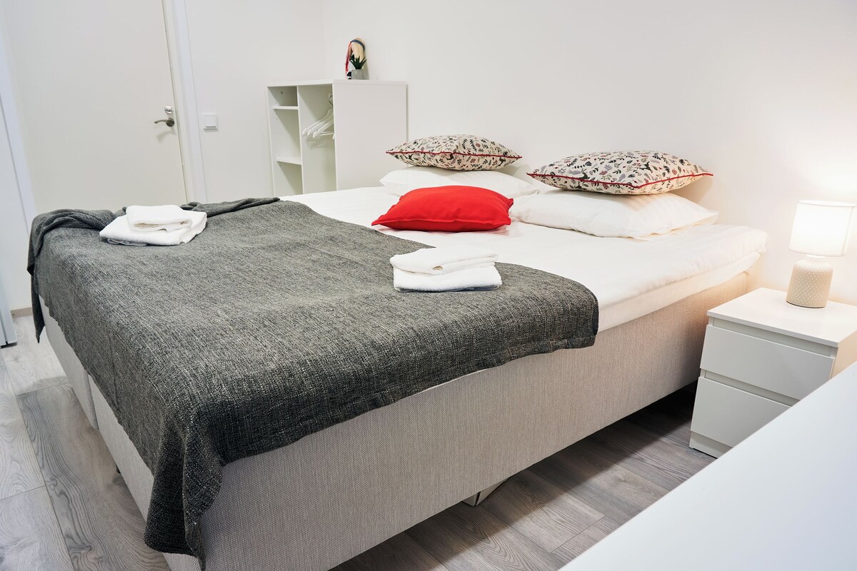 Una cama bien hecha con una manta gris, almohadas blancas y una almohada decorativa roja. Hay una mesita de noche blanca con una lámpara y un pequeño estante encima.