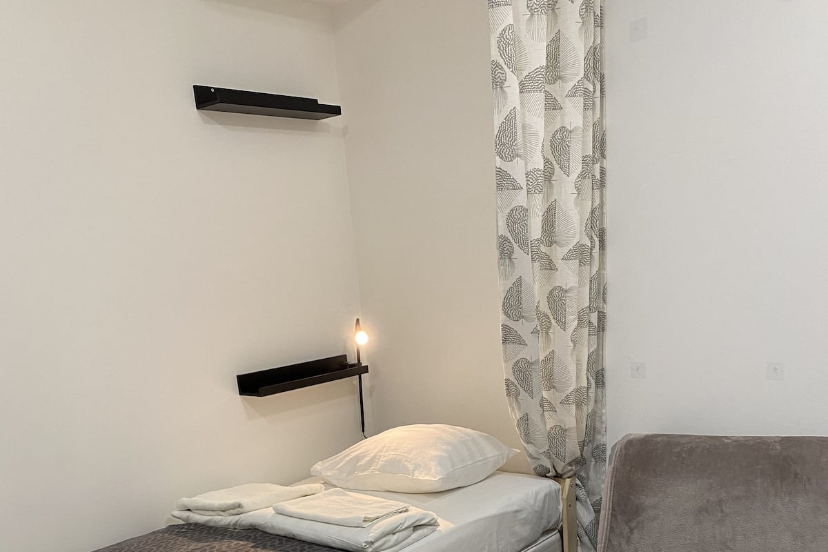 A small, compact room with a single bed, a bedside table, a chair, and a curtain.
