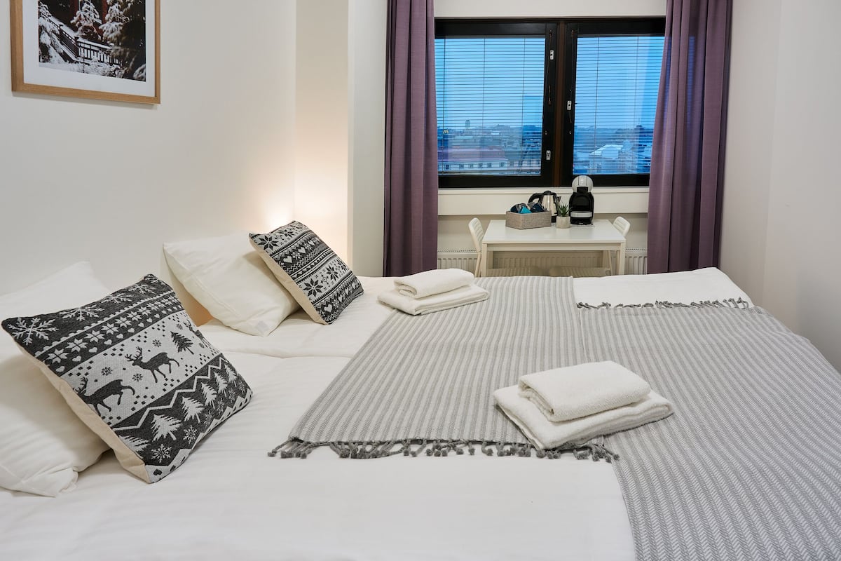 A neatly made bed with patterned pillows, a striped blanket, and folded towels. A small table with a kettle and a basket is visible in the background.