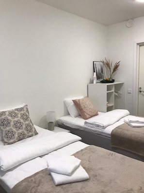 A hotel room with two beds, a bedside table, a nightstand, a white console table with decorative items, and a door. - InnTourist Hostel (Helsinki)