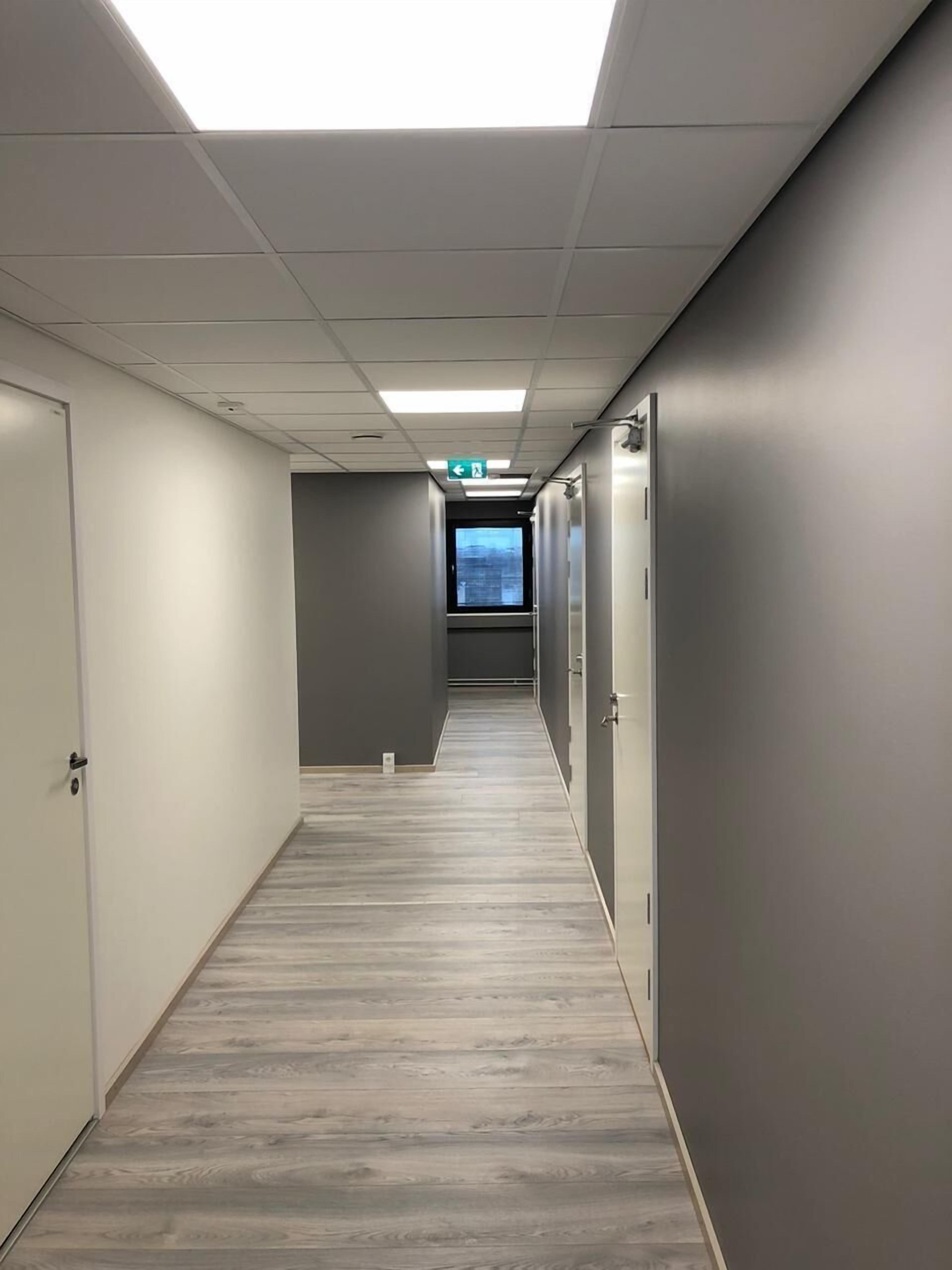 A corridor with grey walls, wooden flooring, and a ceiling with fluorescent lights.