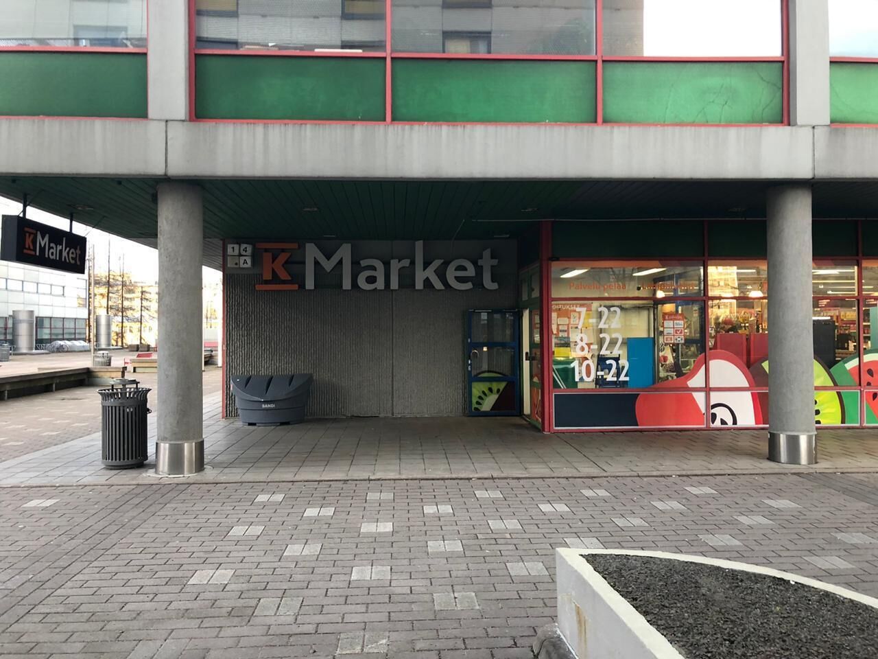 Una tienda K Market con fachada moderna, ventanas grandes y un bote de basura a la vista afuera.