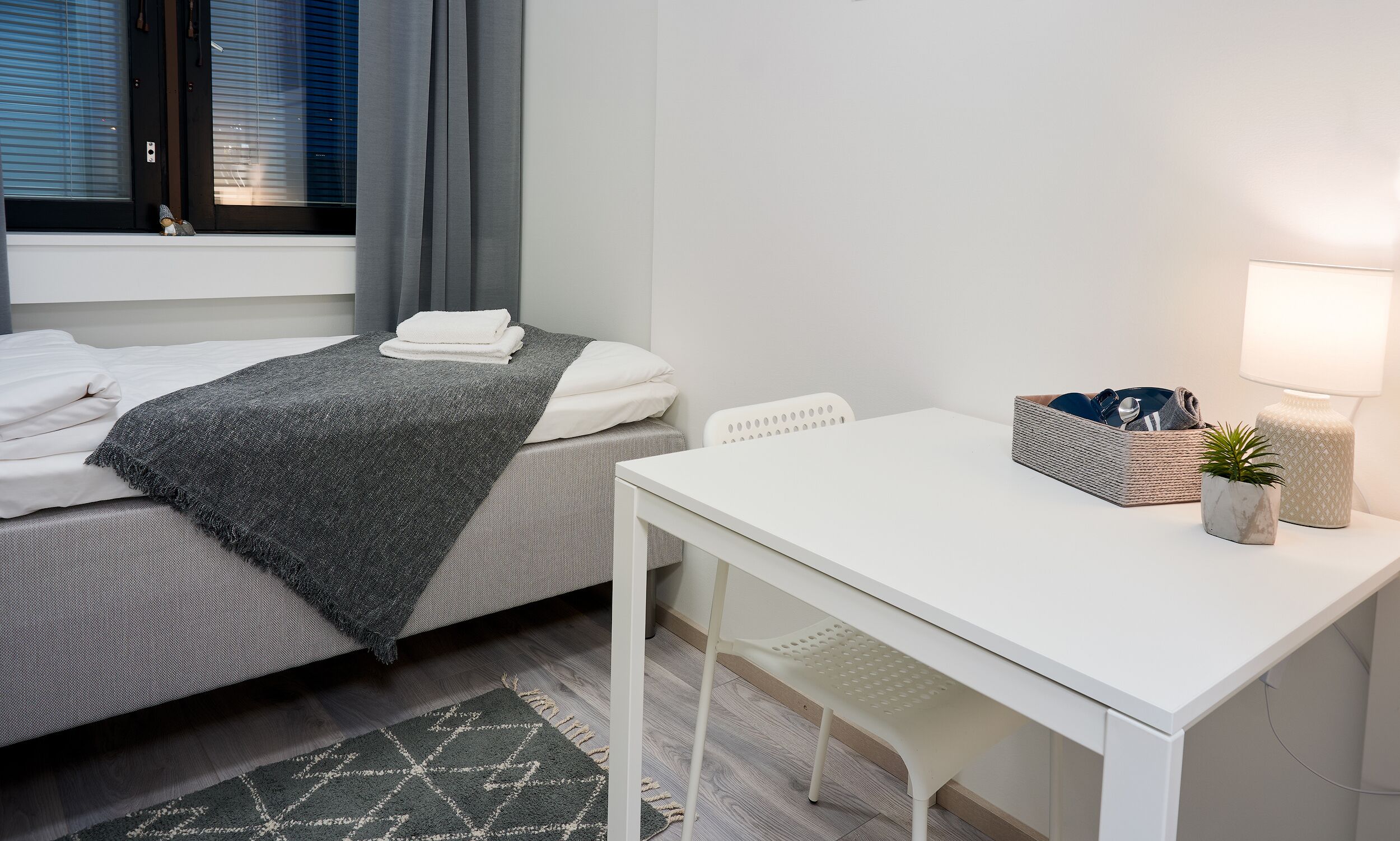 Un dormitorio ordenado con cama, mesita de noche, lámpara y una planta pequeña.