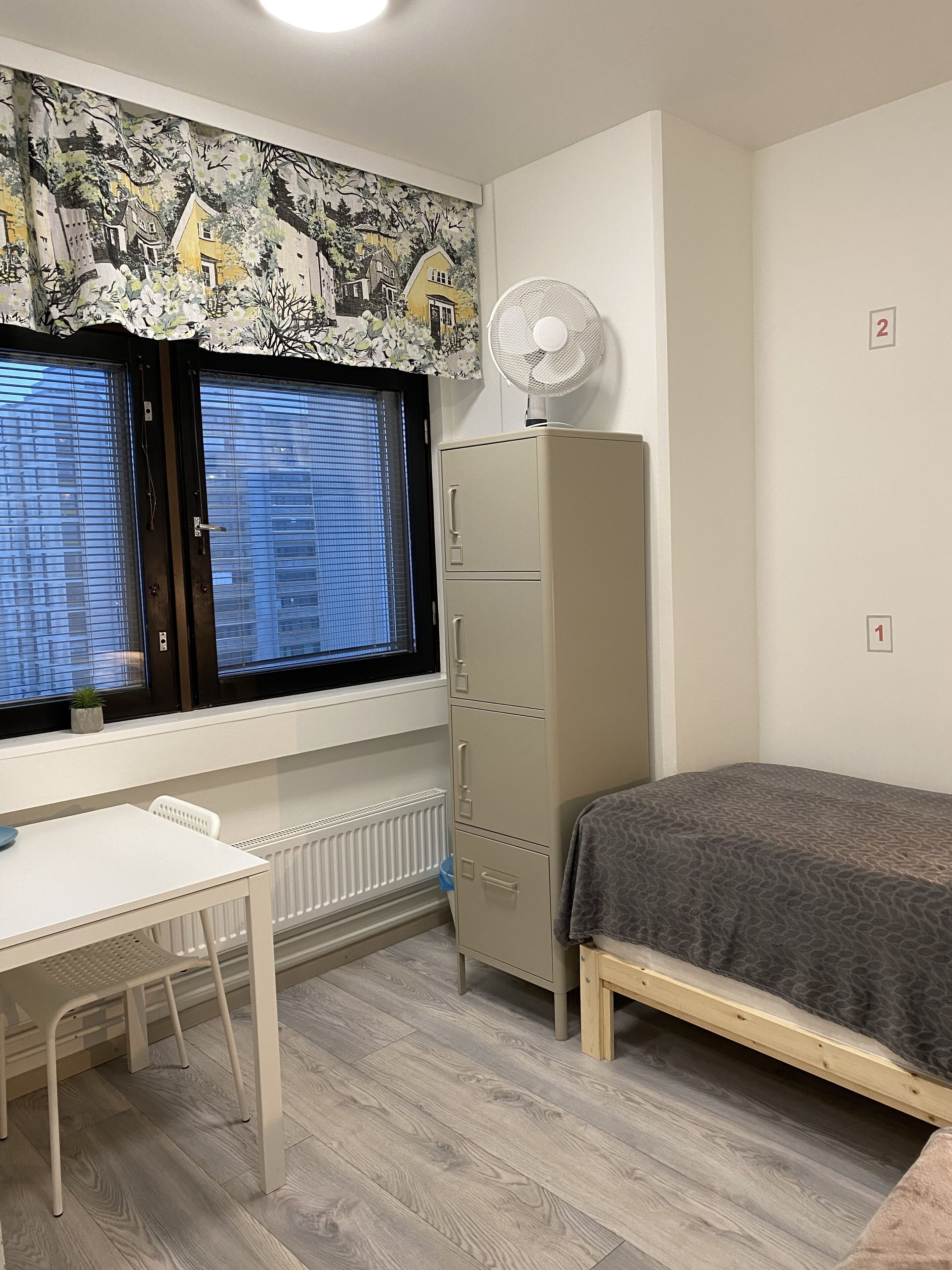 Una habitación pequeña y bien iluminada con ventana, escritorio, cama y radiador.