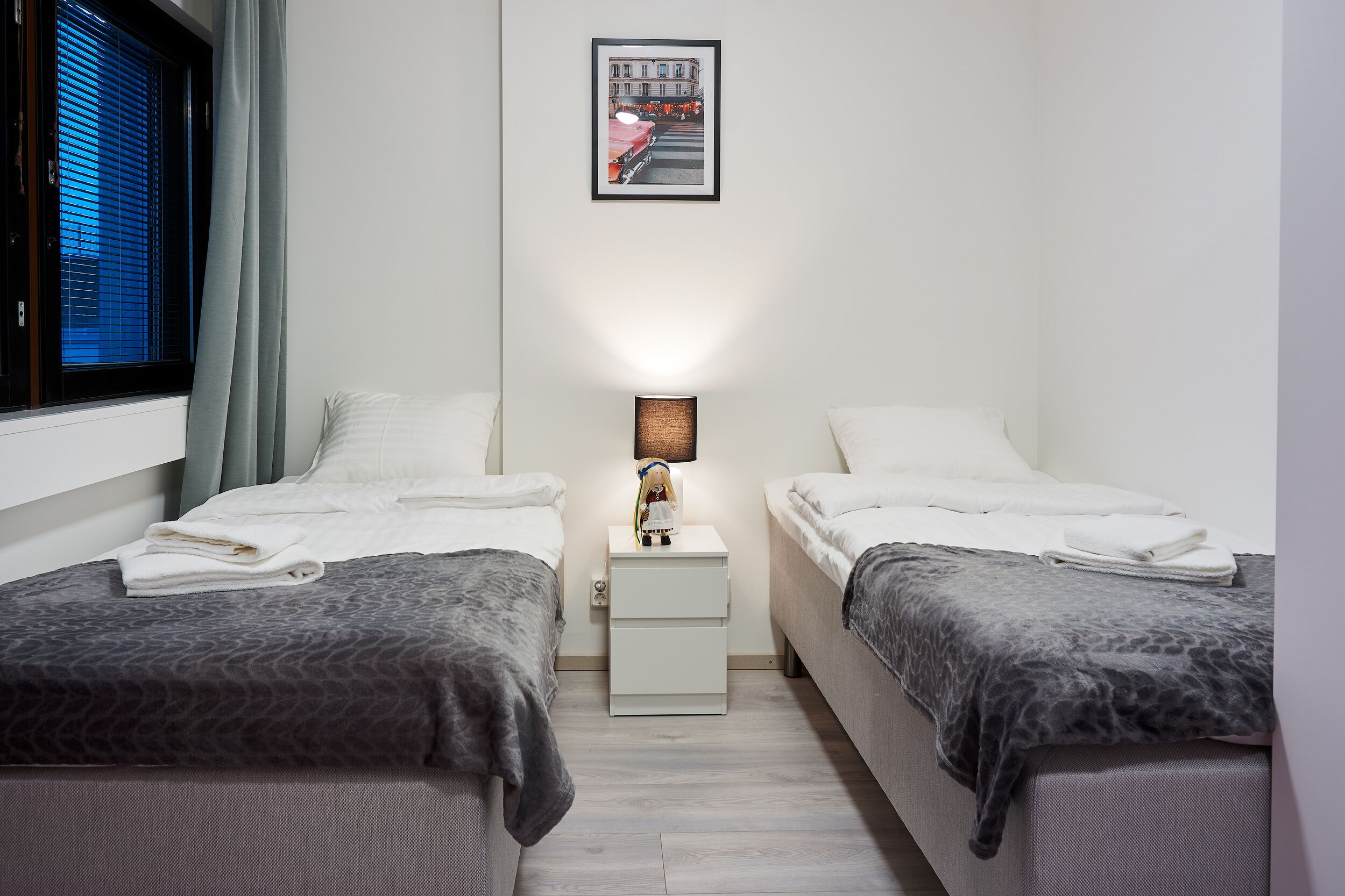 Un dormitorio doble con dos camas, una mesita de noche con lámpara, una ventana con persianas y un cuadro en la pared.
