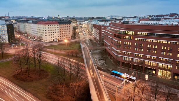 Un immeuble à plusieurs étages avec une façade rouge, un parking et une route où circule un autobus.