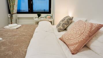 Una cama bien tendida con almohadillas estampadas, una mesita de noche con una planta y una ventana con persianas.