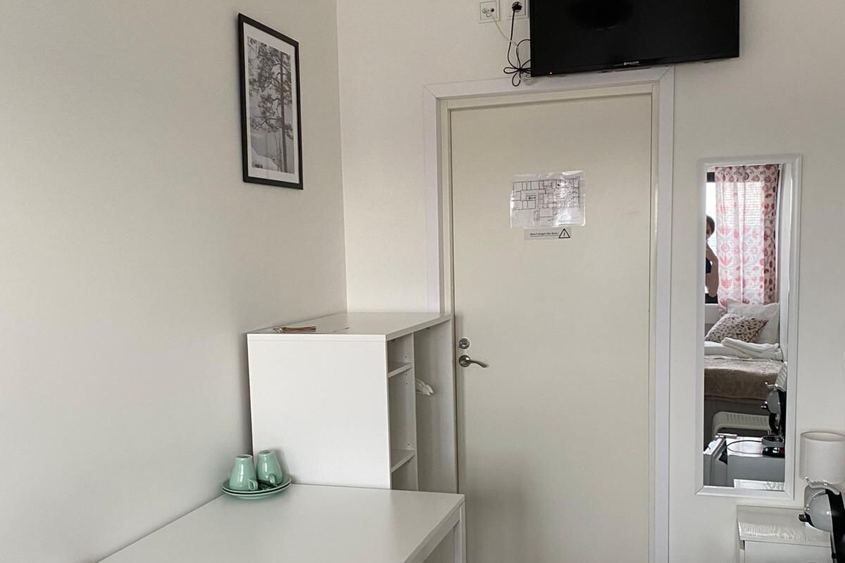 Una habitación pequeña y limpia con un escritorio blanco, dos sillas, una cama con colcha estampada en rosa y blanco, un televisor montado en la pared y una puerta con un cartel.