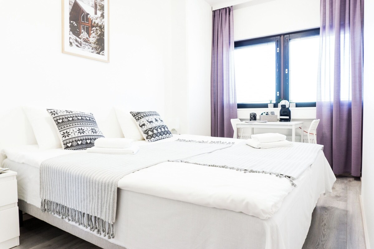 Un dormitorio ordenado con una cama, almohadas, una mesita de noche y una ventana con cortinas.