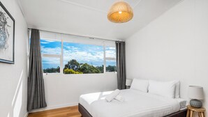 A bedroom with a bed, a window with curtains, a wooden floor, and a hanging light fixture.