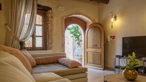 A cozy living room with a large sofa, a small round table with a vase, a television, and a wooden door leading to an outdoor area. - Villa with garden, inneryard and terrace with a view to Talean Mountains (Melidoni Rethymni)