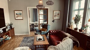 A living room with a leather sofa, a wooden coffee table, a fur-covered ottoman, and a patterned rug. There are two framed artworks on the walls, a chandelier, and a window with curtains. - Agardhs Pensionat (Sveg)
