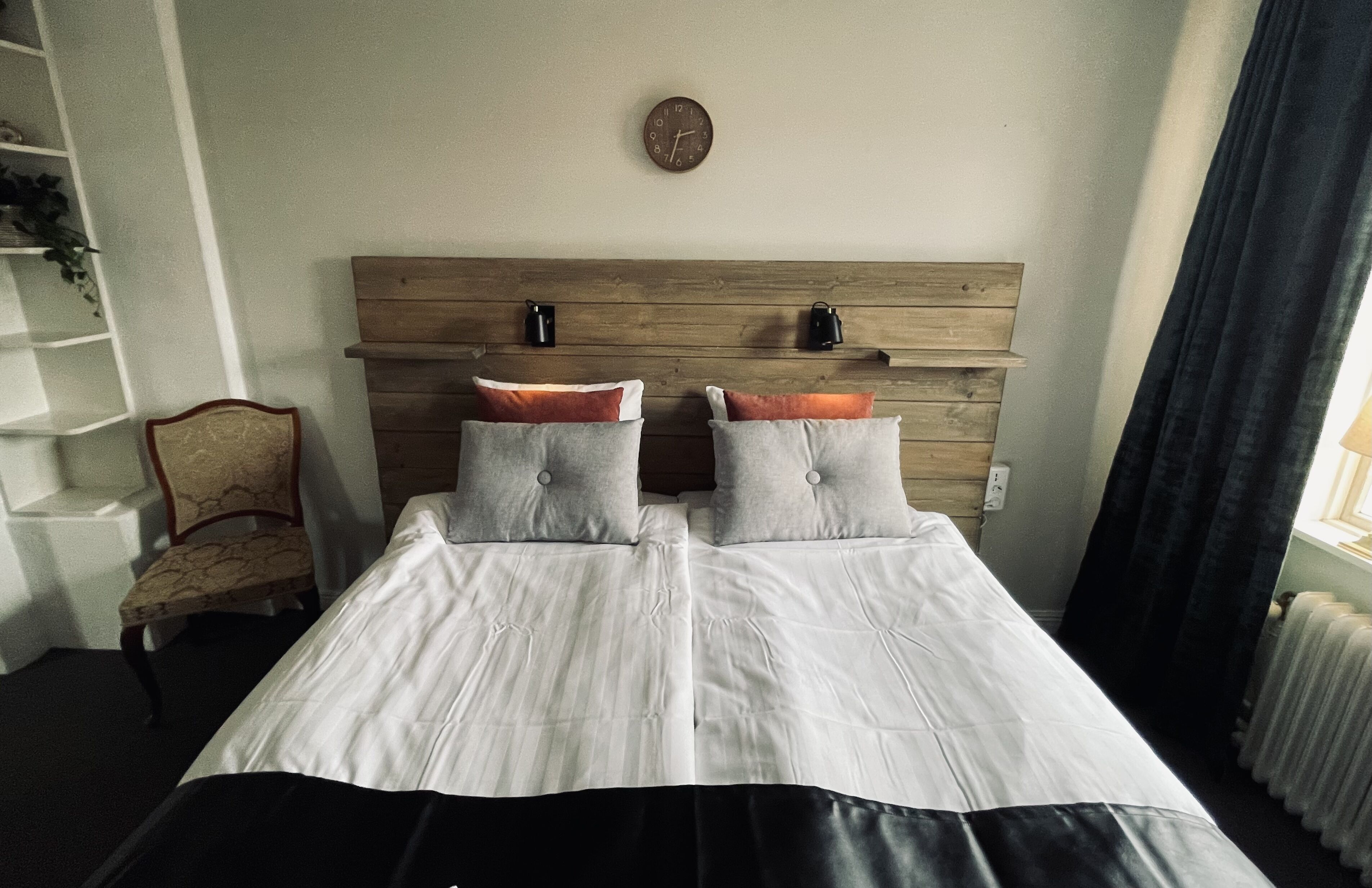 A double bed with two pillows, a wooden headboard, a wall clock, a radiator, and a chair.