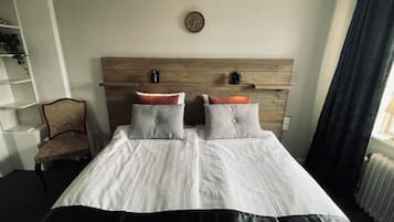 Una cama doble con dos almohadas, cabecera de madera, un reloj de pared, un radiador y una silla.