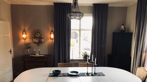 A dining area with a round table, chairs, wall-mounted light fixtures, and a window with curtains. - Agardhs Pensionat (Sveg)