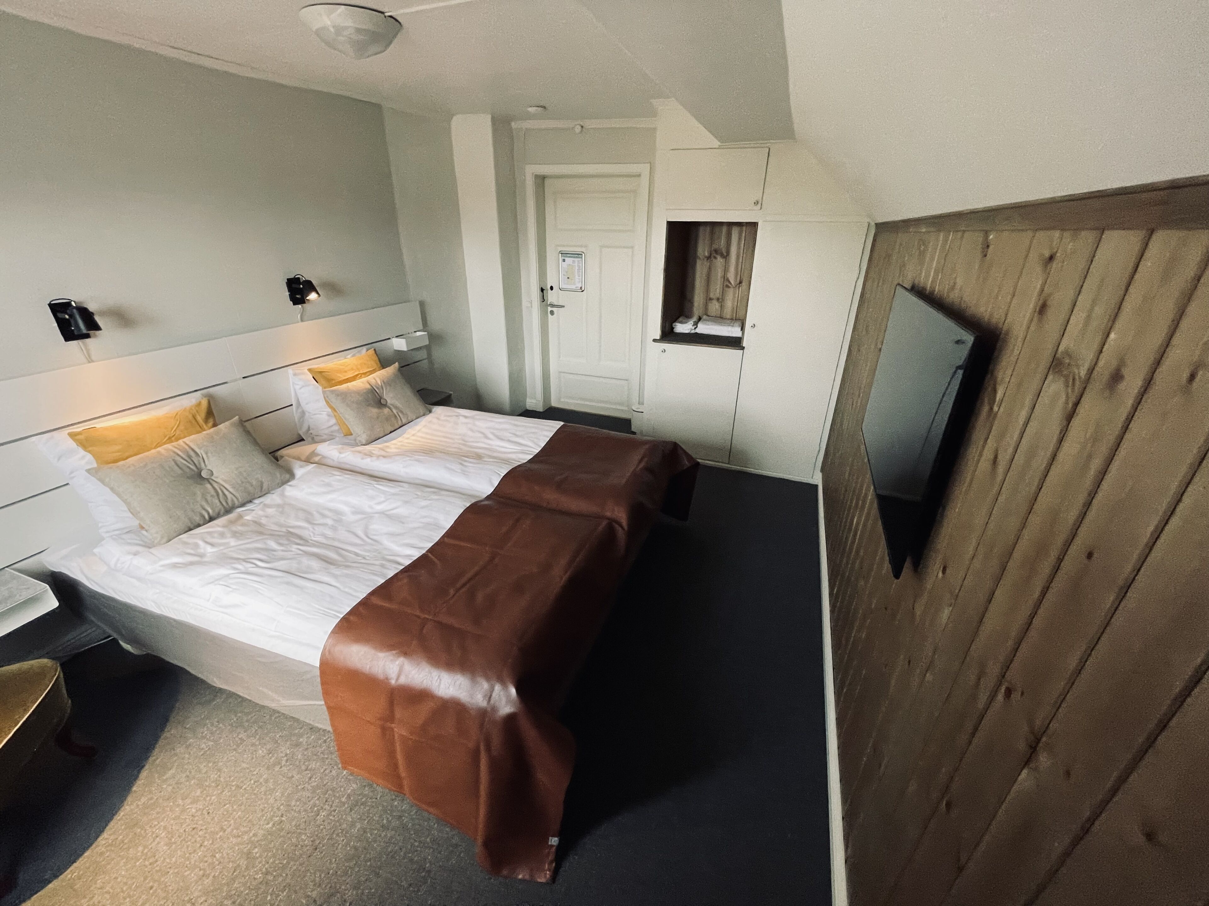 A hotel room with a bed, bedside tables, a TV mounted on the wall, and a wooden panel on one side.