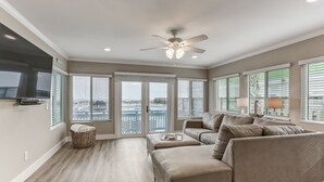 A spacious living room with a large beige sectional sofa, a ceiling fan, and a view through large windows to the outside. - Oceanfront Beach House with Spacious Backyard by RedAwning (Fernandina Beach)